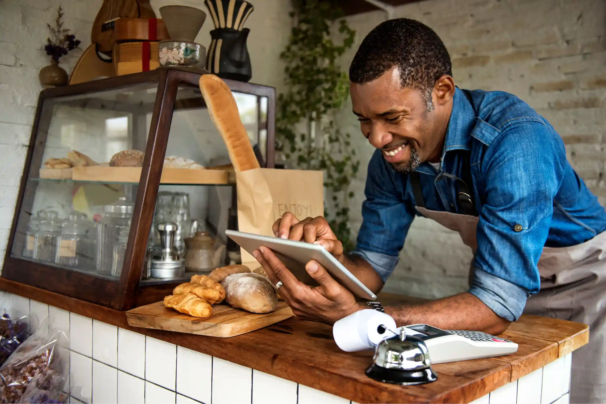 A smiling baker in an apron leaning on the countertop while using a tablet.